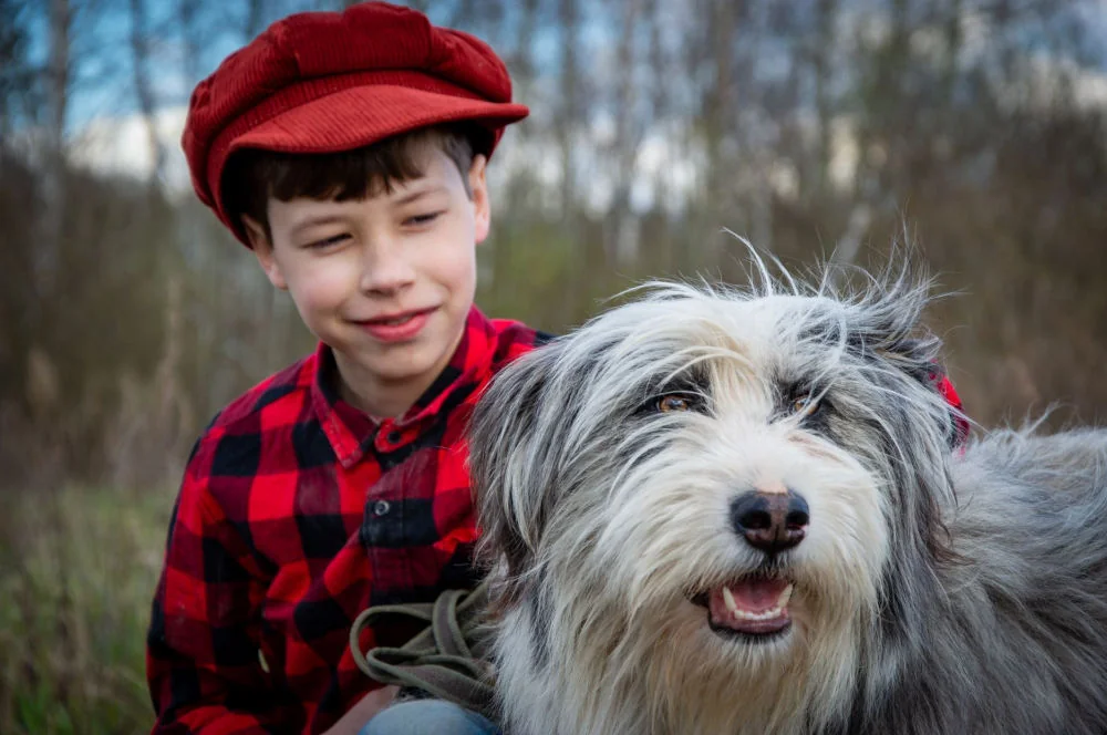 Portret psa o długiej, gęstej sierści z wyraźną brodą i włosami nad oczami; obok siedzi chłopiec na zewnątrz.